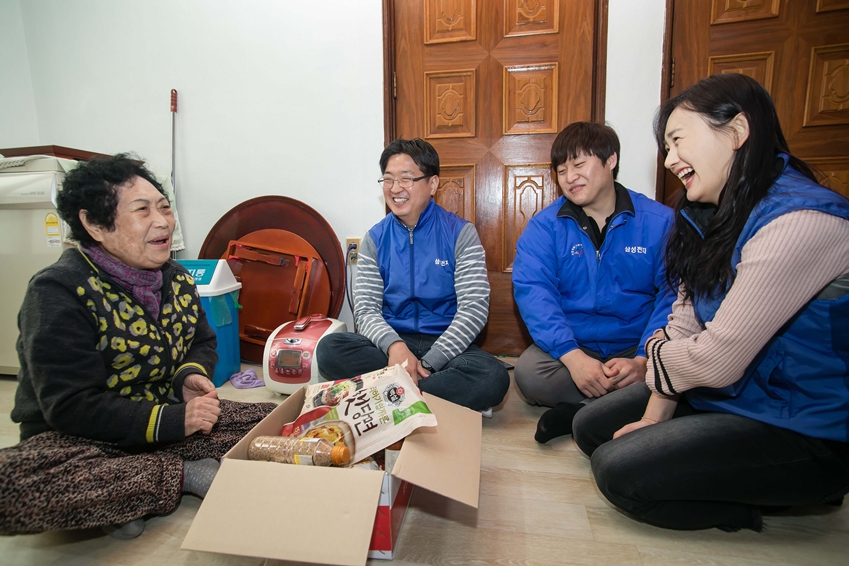 임직원 봉사자들이 어르신의 집을 방문해 부식품 세트를 전달하고 웃으며 대화하고 있다. 